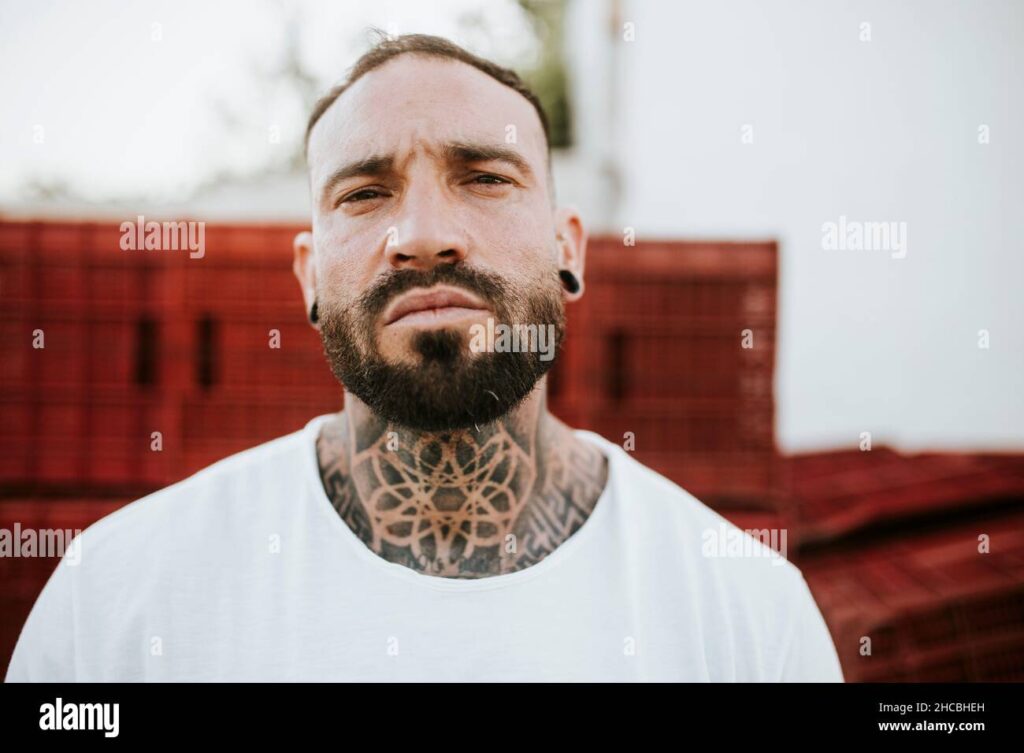Homme barbu avec un tatouage stylé sur le cou et des oreilles percées, représentant une idée tendance pour un tatouage masculin.